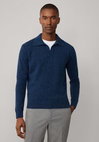 Marineblauwe gebreide trui met korte kraag, textuurpatroon en geribde zoom; gecombineerd met lichtgrijze broeken.