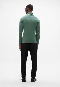 Grünes langärmliges Polohemd mit hohem Kragen, kombiniert mit schwarzen Hosen. Der Stoff wirkt glatt, passgenau und hat ein sauberes Design.