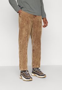Brązowe spodnie w korduroy z prostymi nogawkami, z wyraźnymi pionowymi szwami. W połączeniu z ciemnymi butami sportowymi z siateczkowymi i gumowymi akcentami.
