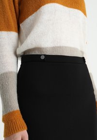 Strickpullover mit orange, weiß und grauen Streifen, kombiniert mit einem hochgeschnittenen schwarzen Rock aus glattem Material mit einer Knopfdetails.