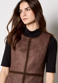 Brązowa kamizelka z sztucznego zamszu z dekoracyjnym haftem, z okrągłym dekoltem i bezrękawowym krojem, noszona na czarną bluzkę z długim rękawem.