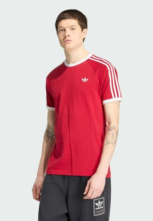 Jonge man draagt een rood Adidas T-shirt met witte strepen op de mouwen en zwarte Adidas shorts met logopatch op het linkerbeen.