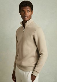 Beige ribbet pullover med kvart-lynlås, blød tekstur og tætsiddende manchetter. Stylet med creamefarvede bukser.
