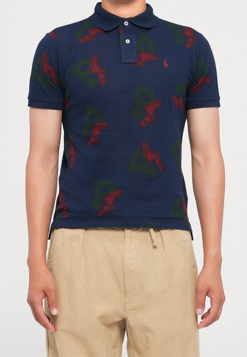 Homme portant un polo bleu marine avec des lettres vertes et des motifs de chaussures ailées rouges, associé à un pantalon beige sur un fond uni.