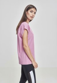 T-shirt di cotone rosa con maniche corte arrotolate e scollo tondo. Presenta una vestibilità rilassata e una texture morbida, abbinata a leggings neri.