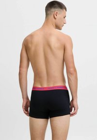 Moški s kratkimi lasmi, oblečen v črne bokserske spodnje hlače z rdečim pasom, označenim z "Jack & Jones," prikazan s hrbtne strani na belem ozadju.