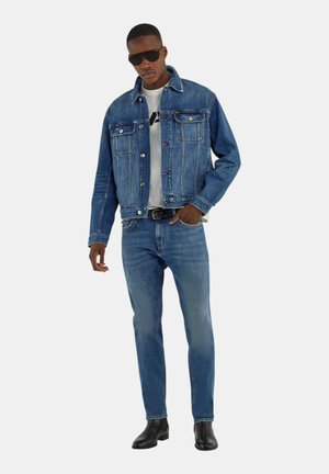 Uomo con occhiali da sole scuri, giacca di jeans blu, maglietta bianca con grafica, jeans blu, cintura nera e scarpe di pelle nere in piedi con una mano in tasca.