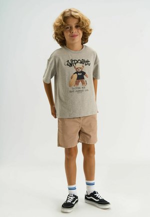 Sivé tričko s grafickým motívom medvedíka a textovým dizajnom. Béžové šortky, čierne Vans s bielymi detailmi a pásikavé biele ponožky. Hladké textúry látky.