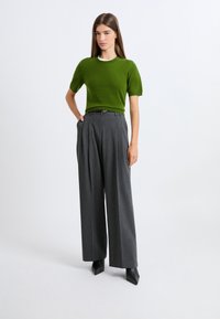 Zielony krótki sweter z dzianiny z krótkim rękawem, szare szerokie spodnie z plisami, czarne szpilki z zaostrzonym czubkiem, noszone z czarnym paskiem na jednolitym tle.