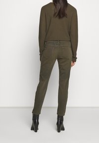 Pantalon vert olive à coupe slim, fabriqué en tissu de coton doux. Dispose de deux poches arrière et de détails de couture subtils. Porté avec des bottines noires à talons.
