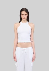 Weißes, geripptes Crop-Top mit hohem Halsausschnitt und ärmellosem Design, kombiniert mit lockeren weißen Hosen mit seitlichen Taschen.
