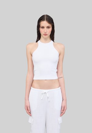 Weißes, geripptes Crop-Top mit hohem Halsausschnitt und ärmellosem Design, kombiniert mit lockeren weißen Hosen mit seitlichen Taschen.