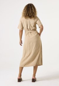 Beige Midi-Kleid mit Gürtel, kurzen Ärmeln, feinen Falten am Rücken und fließender Silhouette, kombiniert mit braunen Absatzsandalen.