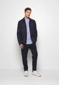 Navy jacket with large front pockets over a blue and white striped shirt, paired with dark pants and white sneakers. Minimalist design.