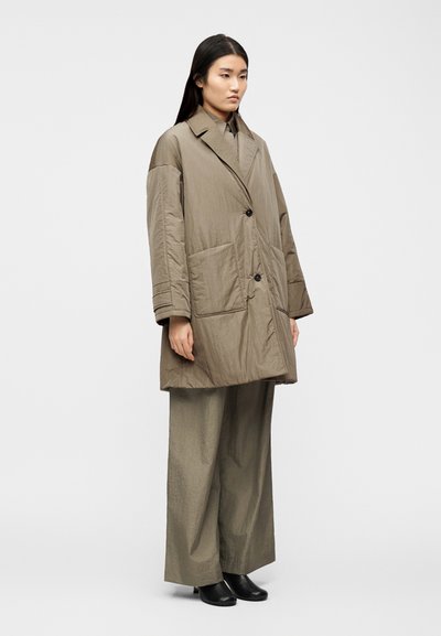 Γυναίκα που στέκεται φορώντας υπερμεγέθη παλτό σε απόχρωση ταουπ και ασορτί παντελόνι με φαρδιά μπατζάκια και μαύρα μποτάκια στους αστραγάλους, σε απλό φόντο.