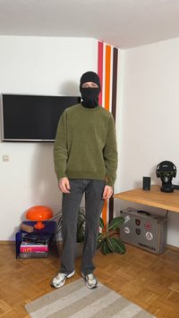 Suéter verde oscuro, pantalones grises, mascarilla negra y zapatillas. Habitación con una lámpara naranja, una planta y un televisor en la pared.