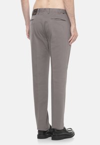 Uomo che indossa pantaloni grigi e scarpe nere, visto da dietro, in piedi di fronte a uno sfondo bianco semplice.