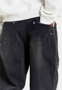 Zwarte denim jeans met een relaxed fit, voorzien van achterzakken en metalen knoopdetails. De stof heeft een zachte, licht versleten textuur.