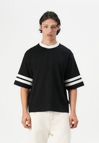 Sort oversized t-shirt med hvid rund halsudskæring og vandrette hvide striber på ærmerne. Fremstillet af bomuld. Har en lige kant.