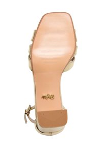 Sole di sandalo con tacco alto beige con superficie liscia e leggermente texture, marchio in rilievo e un accento dorato vicino al cinturino alla caviglia.