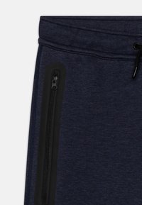 Marineblaue Jogger aus weichem Material, mit einer schwarzen Reißverschlusstasche an der Seite und einem elastischen Bund mit verstellbarem Kordelzug.