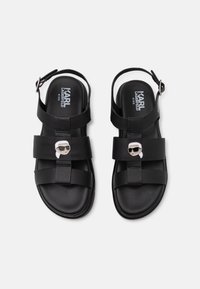 Mustat sandaalit, joissa on kaksi leveää hihnaa ja säädettävä nilkkahihna. Keskihihnassa on hopeinen graafinen koriste. Vaahto- ja teksturoitu pohja.