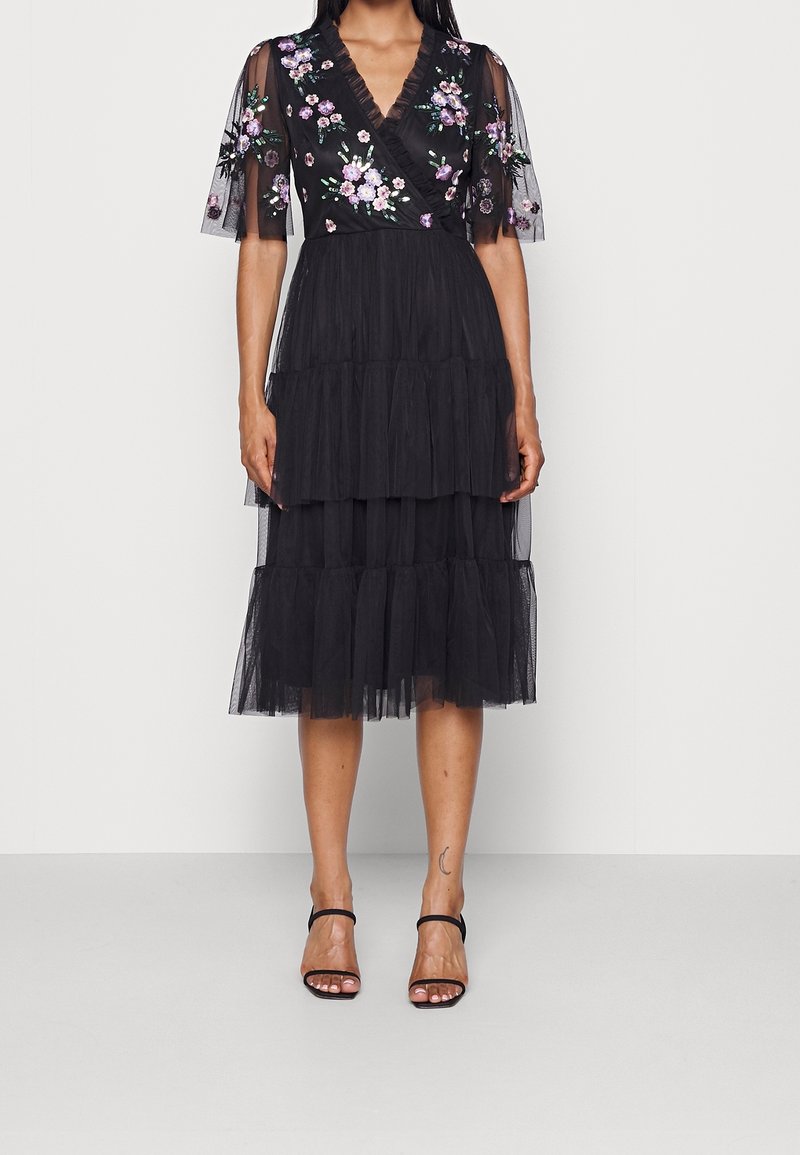 Vestido de tul negro con un diseño en capas, bordado floral en el escote y las mangas, y un corte en V. Se ven tacones negros de tiras.