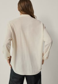 Cream-colored long-sleeve shirt with a collar, smooth texture, and slightly curved hem. Features rolled-up sleeves and a minimalist design.