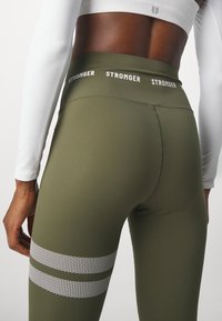 Leggings vert olive avec une taille haute, ornés de texte blanc "STRONGER" et de rayures argentées près des genoux. Matériau lisse et extensible.