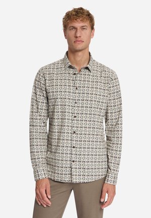 Blonde man die een beige overhemd met knopen draagt met een bruin-wit geometrisch patroon en een bruine broek, staand tegen een effen achtergrond.