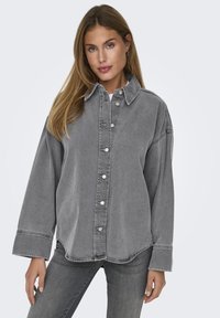 Camicia in denim grigio con bottoni frontali, grande colletto e vestibilità ampia. Presenta accessori in metallo argentato e maniche lunghe. Stile casual e testurizzato.