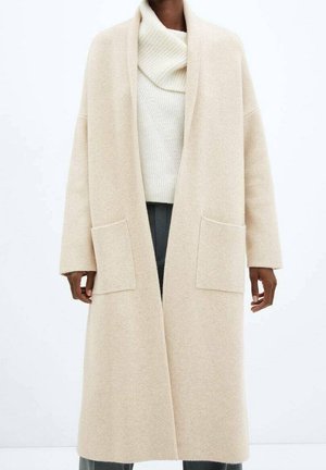 Personne portant un long manteau beige ouvert à l'avant avec de grandes poches frontales, par-dessus un pull à col roulé blanc côtelé et un pantalon foncé.