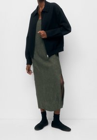 Giacca bomber nera, vestito verde oliva lungo fino al ginocchio con spaccature laterali, calze nere e scarpe basse. Tessuto dalla texture liscia.