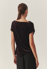 Mørk lilla kortærmet top lavet af blød velour med en bred firkantet halsudskæring og subtile skulderfolder, parret med sorte bukser.