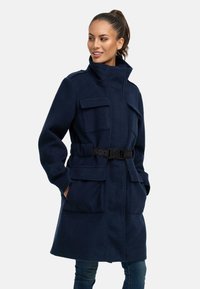 Námornícky modrý fleece kabát s vysokým golierom, opaskom v páse, štyrmi prednými vreckami a prispôsobeným strihom. Hladká textúra s vystavaným dizajnom.