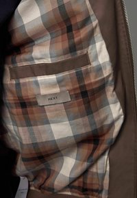 Intérieur d'une veste marron avec un motif à carreaux dans des tons doux de marron, beige et gris. Comprend une poche unique avec une étiquette.