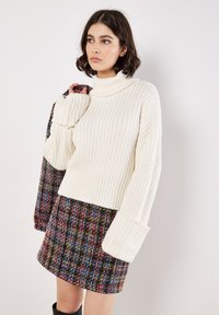 Maglione a collo alto bianco in maglia con dettagli a costine e maniche larghe, abbinato a una minigonna in tweed multicolore con motivo a quadretti.