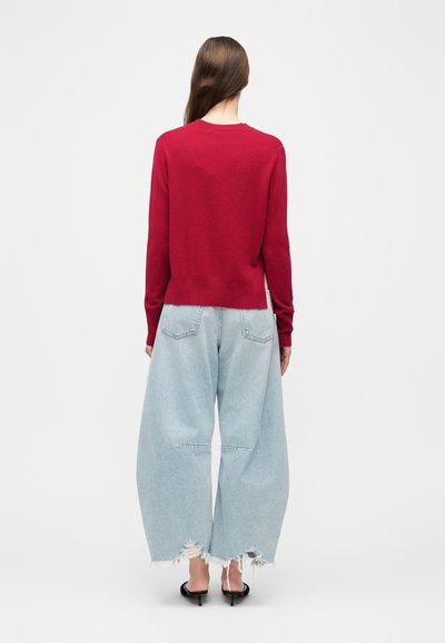 Γυναίκα που στέκεται με την πλάτη προς τα εμπρός, φοράει κόκκινο πουλόβερ, φαρδιά ανοιχτό μπλε σκισμένα τζιν και μαύρα σανδάλια με τακούνι σε λευκό φόντο.