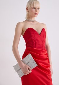 Rochie roșie structurată cu decolteu în formă de inimă și detalii plissate, asortată cu o poșetă cu strasuri argintii. Material din satin, textură fină.
