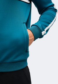 Personne portant un sweat-shirt sarcelle avec une bande blanche sur la manche, main dans la poche avant, debout devant un fond clair.