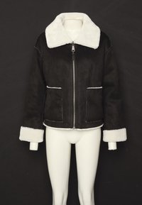 Svart jacka med vit konstpäls på kragen och ärmsluten, uppställd på en mannequin mot en mörk bakgrund, med dragkedja fram och fickor.