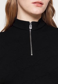 Haut zippé noir avec col haut, présentant un motif texturé et un accent de fermeture éclair en argent. Tissu doux avec une coupe ajustée.
