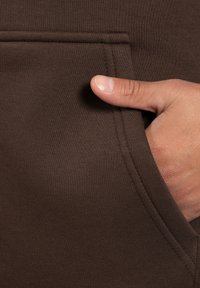 Tejido de sudadera marrón con un bolsillo cosido. Una mano está metida en el bolsillo, mostrando solo el pulgar, destacando la suavidad de la textura y la simplicidad.