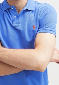 Persona con una polo blu a maniche corte con un logo arancione ricamato sul petto, braccia incrociate sul torso.