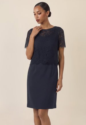 Frau mit dunklen Haaren im Dutt trägt ein marineblaues knielanges Kleid mit Spitzenbesatz am Oberkörper und kurzen Ärmeln, steht vor einem neutralen Hintergrund.
