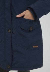 Tamsiai mėlynas izoliuotas švarkas su sagų uždarymu, didelėmis priekinėmis kišenėmis ir logotipo pleistru. Matoma tekstūruota medžiaga ir raukšlėtos rankogaliai.