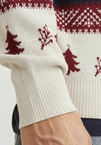 Pull en tricot avec une base crème, ornée de motifs géométriques et d'un arbre bordeaux, des poignets côtelés et une texture douce.