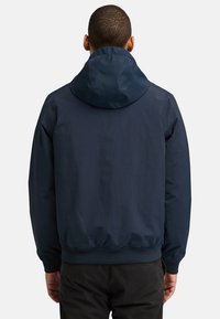 Marinblå jacka med huva, som har en slät yta och elastiska ärmslut. Bakåtblick visar en avslappnad passform och rynkad nederkant.