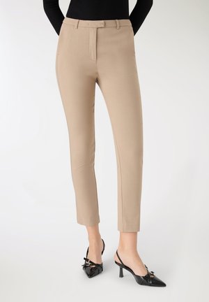 Pantaloni beige aderenti con una texture liscia e una parte anteriore piatta, dotati di una cintura con passanti e silhouette slim, abbinati a tacchi neri.