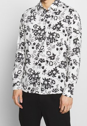 Mężczyzna ubrany w białą koszulę na guziki z długim rękawem w czarne kwiatowe wzory, połączoną z czarnymi spodniami, widoczny od szyi do bioder.